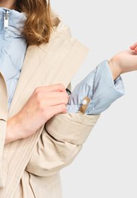 Femme portant un trench-coat beige ajustant la manche d'une veste intérieure bleu clair avec un bouton-pression sur un fond gris.