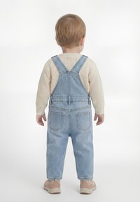Mazs bērns ar gaiši brūniem matiem, ģērbies zilās džinsa kombinezonā, krēmkrāsas dzijas džemperī un bēšās kurpēs, stāvot ar muguru pret baltu fonu.
