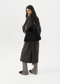 Donna in lungo cappotto di pelle nera e pantaloni grigi, che porta una grande borsa a tracolla in suede nera, mentre guarda oltre la spalla contro uno sfondo bianco.