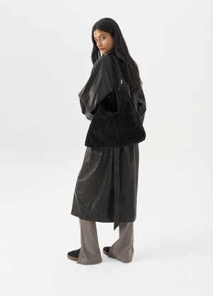 Donna in lungo cappotto di pelle nera e pantaloni grigi, che porta una grande borsa a tracolla in suede nera, mentre guarda oltre la spalla contro uno sfondo bianco.