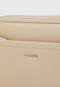 Bež torba od umjetne kože s glatkom teksturom, sadrži srebrne detalje i vidljivi Calvin Klein logo. Jednostavan, elegantan dizajn s detaljima šivanja.