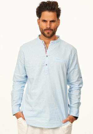 Mann mit Bart, der ein hellblaues Hemd mit Stehkragen und weiße Hose trägt, steht mit den Händen in den Taschen vor einem weißen Hintergrund.