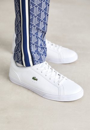 Zapatillas blancas con una textura suave, que presentan un logo de cocodrilo verde, cordones planos y pequeños orificios de ventilación en el lateral.