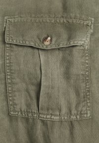 Tessuto verde oliva con una tasca frontale dotata di chiusura a bottone. La texture è leggermente stropicciata, suggerendo un materiale leggero.