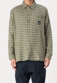 Grön rutigt skjorta med knäppning fram, krage och bröstficka med en logotagg. Tillverkad av textil med synlig sömnad.