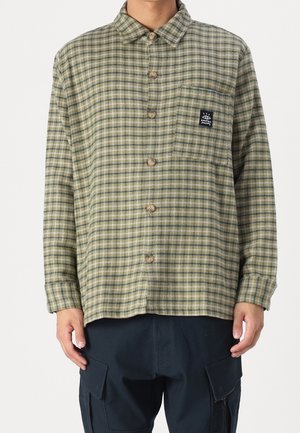 Camisa de cuadros verdes con botonera en la parte delantera, cuello y un bolsillo en el pecho que presenta una etiqueta de logo. Fabricada con tela texturizada y costuras visibles.