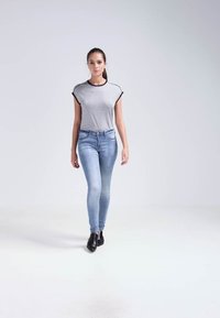 Camiseta de rayas grises con ribete negro, combinada con jeans ajustados de color azul claro; la modelo lleva botas negras hasta el tobillo, de pie sobre un fondo blanco.