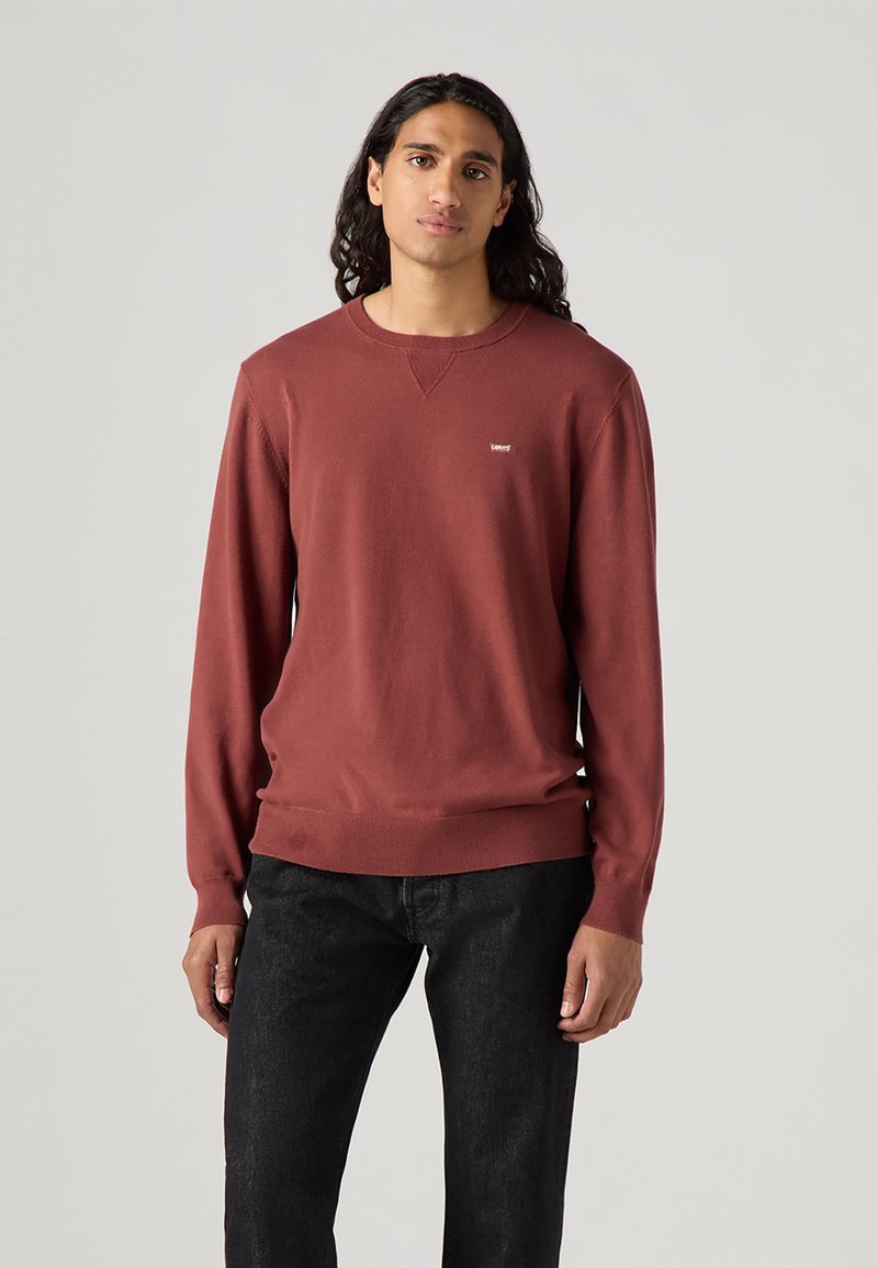 Un pull-over crewneck bordeaux avec des manches longues, fabriqué en tissu doux, présentant un petit logo sur la poitrine et des bords côtelés aux poignets et à l'ourlet.