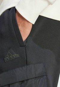 Black athletic pants featuring a textured fabric, a prominent logo, and a contrasting pocket design. A light-colored sweatshirt sleeve is visible.