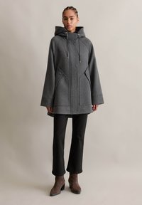 Manteau en laine grise avec une grande capuche, un cordon de serrage et des boutons sur le devant. Il a une coupe décontractée avec des poches latérales et une texture lisse.