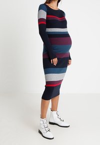 Hladký pletený těhotenský šaty s horizontálními pruhy v námořnické modři, červené, šedé a tyrkysové. Přiléhavé dlouhé rukávy a délka do půli lýtek. V kombinaci s bílými botami na přezku.