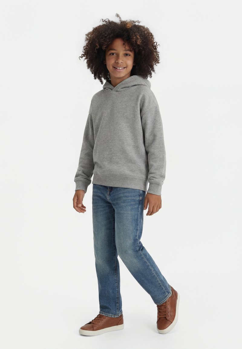 Dijete s kovrčavom kosom koje nosi sivu hoodicu, plave traperice i smeđe tenisice, stojeći s jednom nogom blago podignutom.