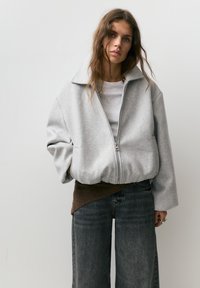 Młoda kobieta z długimi włosami, ubrana w za dużą, szarą kurtkę na zamek, szary t-shirt, ciemne szerokie dżinsy oraz brązową warstwę dzianiny pod spodem.