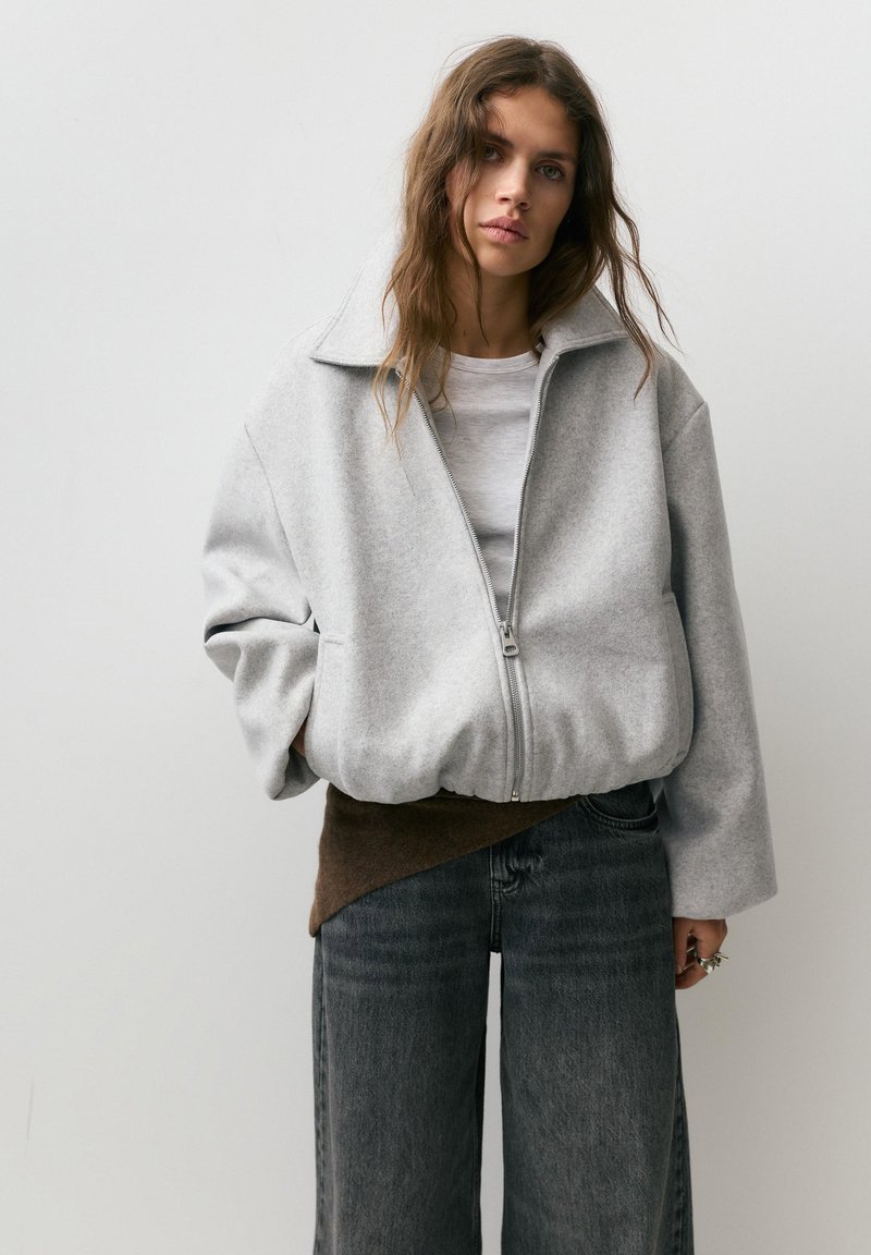 Młoda kobieta z długimi włosami, ubrana w za dużą, szarą kurtkę na zamek, szary t-shirt, ciemne szerokie dżinsy oraz brązową warstwę dzianiny pod spodem.