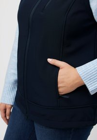 Navyblauer Softshell-Weste mit durchgehendem Reißverschluss, Taschen an der Seite und passgenauer Form, getragen über einem hellblauen Pullover mit gerippten Bündchen.