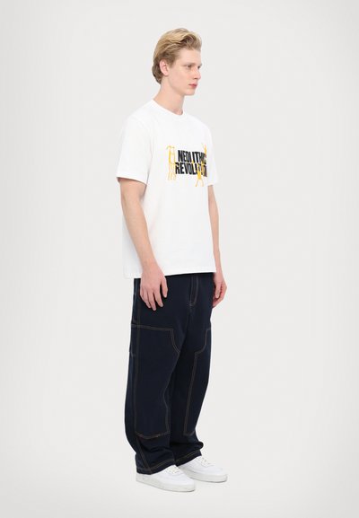 Tricou alb din bumbac cu grafică "REVOLUȚIA NEOLITICĂ" în negru și galben, asortat cu pantaloni largi de culoare bleumarin închis, cu cusături portocalii. Adidași albi.