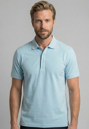 Man met lichtbruin haar en baard, gekleed in een lichtblauw poloshirt met korte mouwen, staand voor een effen grijze achtergrond.