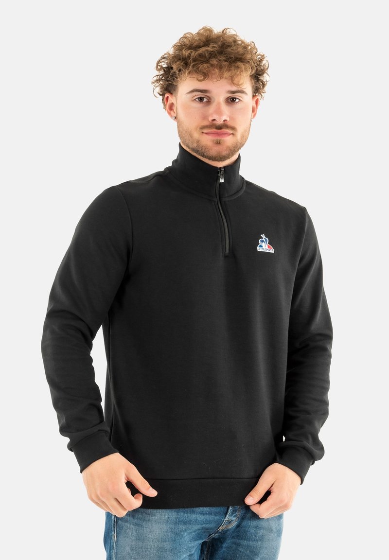 Zwarte sweatshirt met ritssluiting gemaakt van zachte stof, met ribbels aan de mouwen en de onderkant, en een klein gekleurd logo op de linkerkant van de borst.