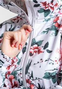 Chaqueta con estampado floral sobre una base blanca, que presenta flores verdes y rojas. El tirador del zipper tiene un acento de rayas multicolores.