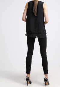 Top nero senza maniche con un orlo inferiore trasparente abbinato a leggings neri aderenti e scarpe col tacco nere, visto da dietro.