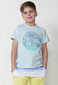 Šviesiai mėlynas medvilninis marškinėliai su apvaliu grafiniu piešiniu, kuriame matomi žali ir mėlyni bangų motyvai, ir užrašas "SURFER LIFE" paryškintomis raidėmis.