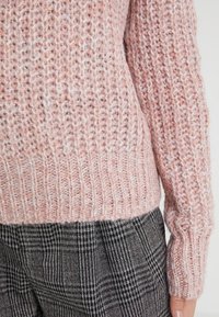 Maglione chunky a maglia grossa di colore rosa chiaro, con polsini e orlo a coste. Indossato sopra pantaloni a quadri neri e grigi. Aspetto testurizzato e accogliente.