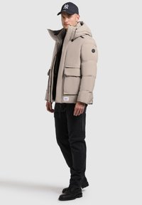 Beige puffer jacket with a hood, featuring two front pockets, black hardware accents, and a logo patch. Worn over a black shirt and jeans.