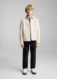 Lichtbeige jack met een knoopsluiting aan de voorkant en twee grote zakken, gedragen over een zwart shirt, gecombineerd met zwarte broek en witte sneakers.