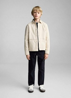Lichtbeige jack met een knoopsluiting aan de voorkant en twee grote zakken, gedragen over een zwart shirt, gecombineerd met zwarte broek en witte sneakers.