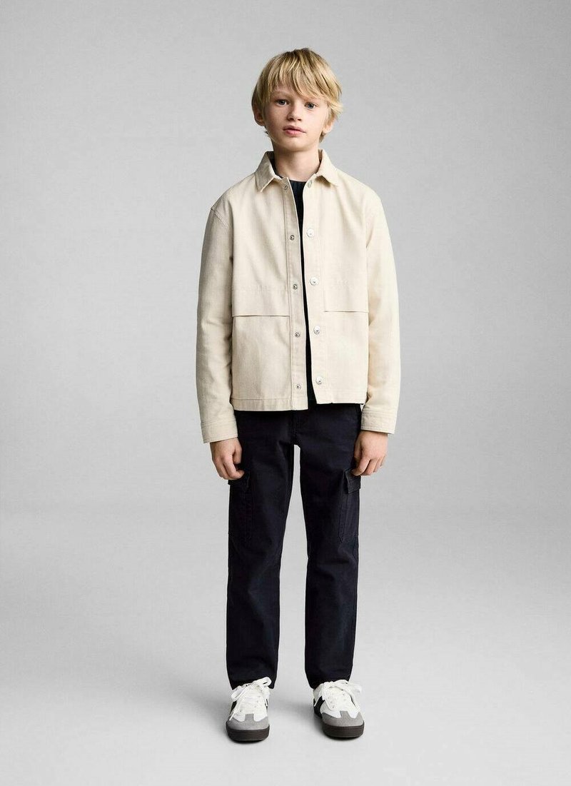 Lichtbeige jack met een knoopsluiting aan de voorkant en twee grote zakken, gedragen over een zwart shirt, gecombineerd met zwarte broek en witte sneakers.