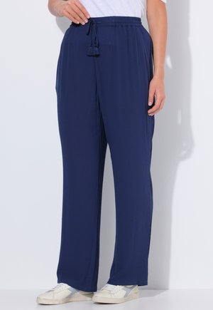 Persona vistiendo pantalones anchos de pierna en azul marino con cintura de cordón y zapatillas blancas, de pie contra un fondo blanco.