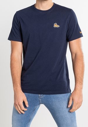 Mann trägt dunkelblaues T-Shirt mit kleinem gelbem Stiefellogo auf der Brust und hellblaue Jeans, steht mit entspannt hängenden Händen an den Seiten.