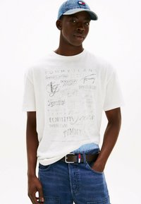 Camiseta de algodón blanca con varios logos en gris de "Tommy Jeans", combinada con shorts de mezclilla oscura y una gorra de mezclilla azul acentuada por una franja roja y blanca.
