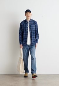 Camisa de cuadros azul y verde sobre una camiseta blanca, jeans azul claro, zapatos beige y una gorra navy. Sostiene una bolsa de tote beige.