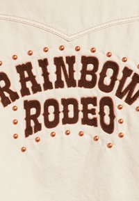 Bavlněná látka s krémovým pozadím zdobí výšivka &quot;RAINBOW RODEO&quot; v tmavě hnědé barvě, doplněná měděnými nýty.