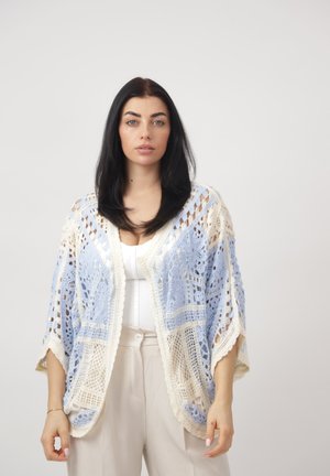 Vrouw met lang zwart haar draagt een lichtblauwe en beige gehaakte sjaal over een witte top en beige broek, staand tegen een effen achtergrond.