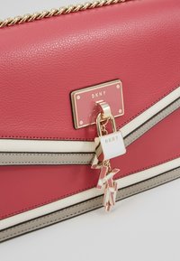 Sac en cuir rose avec une chaîne dorée, présentant une plaque logo DKNY et un charm de serrure décoratif avec des accents de logo et des rayures contrastantes.