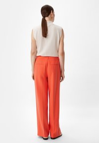 Beżowy top bez rękawów z wysokim kołnierzem; szerokie, jasnopomarańczowe spodnie; gładki materiał; widok z tyłu pokazujący szew tylny; brak widocznych wzorów lub akcentów.