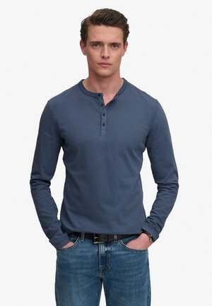 Mężczyzna z krótkimi brązowymi włosami stoi z rękami w kieszeniach, ubrany w niebieską koszulkę henley z długim rękawem, niebieskie dżinsy, czarny pasek i zegarek na nadgarstku.