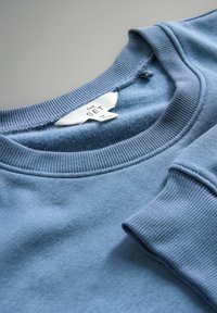 Primo piano di una morbida felpa blu che mostra il collo a costine, il polsino delle maniche e l'etichetta bianca con la taglia e il marchio "THE SET" all'interno del colletto.
