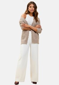 Cárdigan de punto beige con un patrón texturizado, combinado con pantalones anchos blancos y una camiseta sin mangas ajustada blanca. Tacones marrones completan el conjunto.