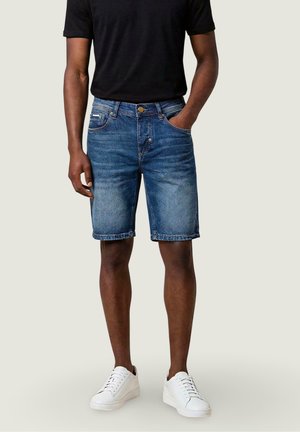 Uomo che indossa pantaloncini di jeans a lavaggio medio, t-shirt nera e scarpe da ginnastica bianche, in piedi con una mano in tasca contro uno sfondo semplice.