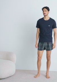 T-shirt azul marinho com um pequeno logótipo, combinado com boxers xadrez pretos. O modelo está de pé sobre um tapete claro ao lado de uma cadeira beige arredondada.