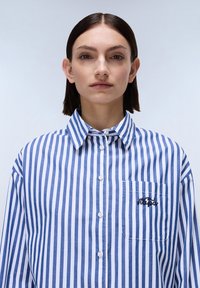 Camicia a righe blu e bianca in cotone, con colletto, tasca frontale e dettaglio ricamato sulla tasca.