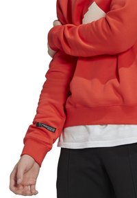 En person som bär en röd sweatshirt över en vit skjorta, justerar sin vänstra ärm med höger hand, svarta byxor synliga från midjan och ner.