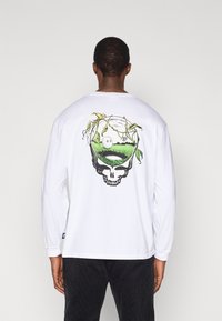 Λευκό πουκάμισο με μακριά μανίκια που διαθέτει γραφικό σχέδιο κρανίου που περιβάλλει μια σκηνή γκολφ, με πράσινο χορτάρι και κληματαριά. Κατασκευασμένο από βαμβάκι, με ελαστικές μανσέτες.