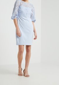 Jasnoniebieska sukienka z koronkowym gorsetem i rękawami do trzech czwartek. Spódnica jest gładka, a do niej noszone są beżowe sandały na blokowym obcasie.