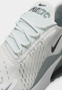 Λευκό αθλητικό παπούτσι Nike Air 270 με μαύρο λογότυπο, αναπνεύσιμο πάνω μέρος από δίχτυ και λευκά κορδόνια, εμφανίζεται σε κοντινή γωνία.