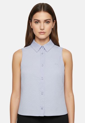 Donna che indossa una camicia senza maniche, azzurro chiaro con una texture e bottoni, collo appuntito, in piedi davanti a uno sfondo semplice.