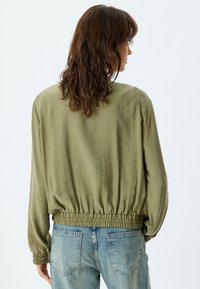 Veste légère vert olive avec un ourlet et des poignets élastiqués, présentant une silhouette décontractée. Associée à un jean en denim bleu clair.